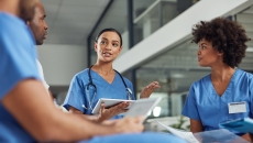 Four healthcare professionals wearing blue scrubs and sitting in a circle with one holding a tablet