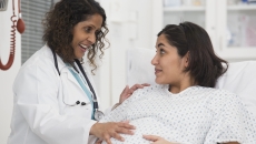 Healthcare worker standing beside a pregnant person lying in bed while both are touching the pregnant person's stomach