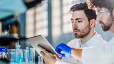 Two people in a laboratory looking at a tablet with beakers behind them