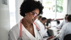 A doctor looking a tablet while a meeting occurs in the background A doctor looking a tablet while a meeting occurs in the background