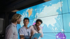 Three people standing in front of a large screen with a map of the world on it