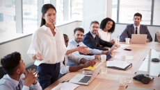 Numerous people in a board room sitting around a table with one person standing up