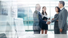 Four people standing in front of a window with two people shaking hands while the two others stand and watch
