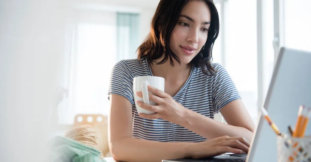 Person in a gray shirt sitting down while holding a coffee mug and looking at a computer Person in a gray shirt sitting down while holding a coffee mug and looking at a computer